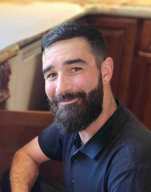Smiling man with well-groomed beard and short dark hair wearing a black polo shirt, sitting indoors.